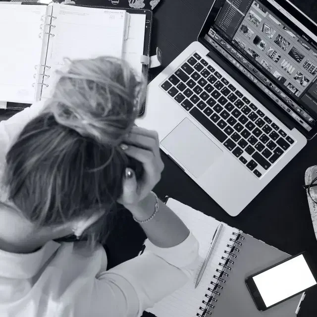 Frau sitzt verzweifelt mit Händen am Kopf über ihren Laptop, Kalender und leeren Blocl.